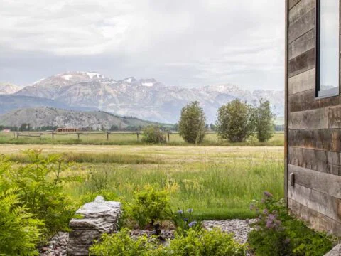 Side view of a rustic house with a small garden in the foreground, overlooking a grassy field and distant mountains under a cloudy sky, embodying Wyoming Home Design that harmonizes with the natural landscape.
