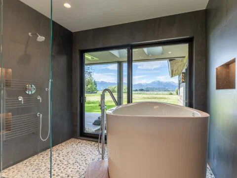 Modern bathroom with a white freestanding tub and glass shower, reflecting lifestyle-driven architecture. Large windows offer a view of mountains and fields, typical of Wyoming home design. Pebble flooring adds texture.