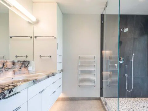 Modern bathroom with a marble countertop, double sink, and large mirror. Tailored Home Solutions feature a glass shower enclosure on the right with tiled walls and a pebble floor, reflecting our dedication to homeowner-centered design.
