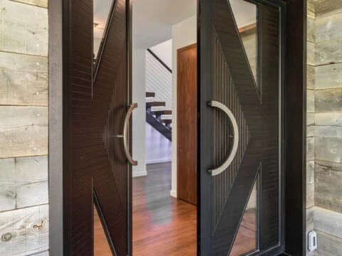 Modern double doors partially open, revealing a hallway with wooden flooring and a staircase inside a house. The doors have large circular handles and geometric design elements, embodying the essence of lifestyle-driven architecture that defines contemporary Wyoming home design.