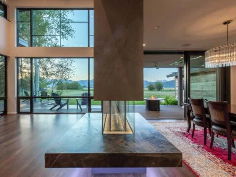 Experience a modern living and dining area by Studio 250 Design, featuring a central fireplace, large windows with scenic views, hardwood floors, and a chandelier above the dining table. Embrace this homeowner-centered design crafted for both style and comfort.