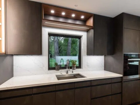 Modern kitchen with dark cabinets, marble countertops, and a stainless steel sink beneath a window. Built-in oven and open shelving with lights enhance the Personalized Architecture, offering tailored home solutions for contemporary living.