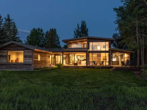 A modern Wyoming Home Design features large glass windows and a wooden exterior, surrounded by trees and a grassy lawn, warmly lit at dusk. This homeowner-centered design creates the perfect tailored home solution for blending elegance with nature.