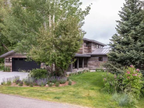 A modern cabin-style house with wooden siding, nestled amid trees and a grassy lawn, embodies lifestyle-driven architecture. It features a garage and a driveway, making it the perfect Wyoming home design.