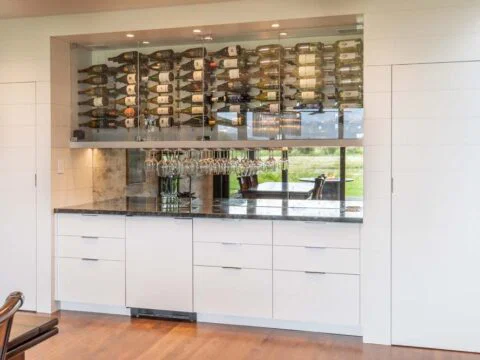 Modern kitchen with a wall-mounted wine rack, storing numerous wine bottles and glassware hanging below. White cabinets with dark countertops complete this Wyoming Home Design, reflecting Lifestyle-Driven Architecture that embodies tailored home solutions.
