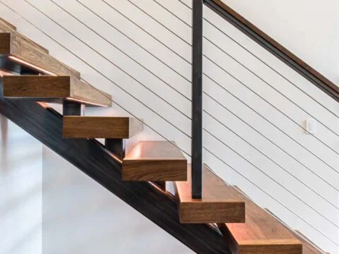 A modern staircase with wooden steps and a steel railing with cable wires, set against a white wall. This Wyoming Home Design showcases open risers and a sleek, minimalist look, emphasizing homeowner-centered design in every detail.